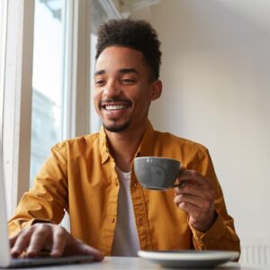 african-american-sitting-table-cafe-working-laptop-wears-yellow-shirt-drinks-aromatic-coffee-communicates-with-his-sister-which-is-far-another-country-enjoys-work-2